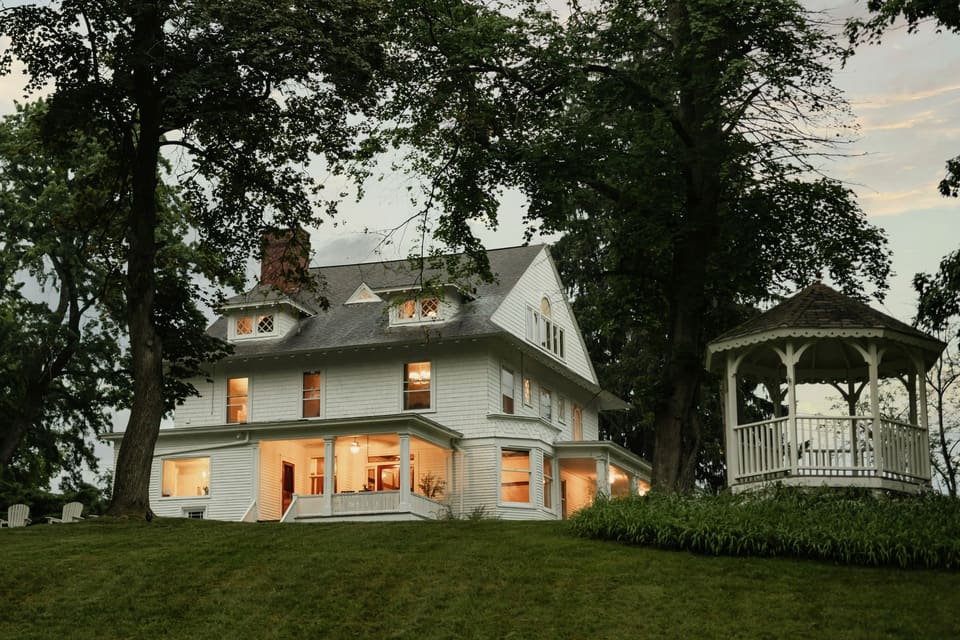 White two-story house at dusk with warm lights on in the windows, a lawn and a gazebo