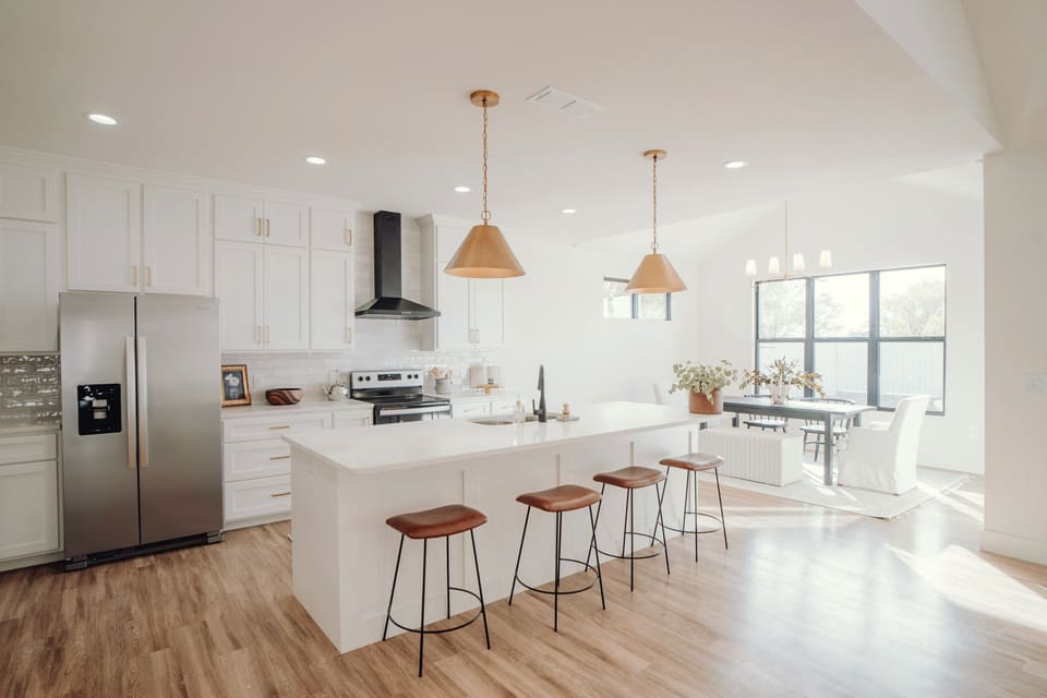 Open kitchen with white cabinets, a long island with four stools and a dining table by the windows