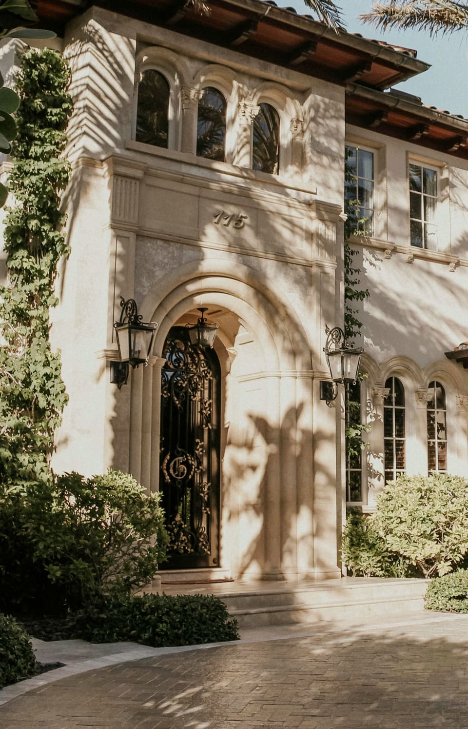 Arched stone entry with a wrought iron door and lanterns on a stucco house, palm shadows across the facade
